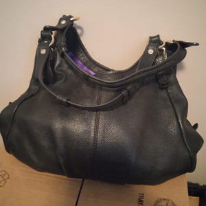 May include: A black leather handbag with a curved shape and a shoulder strap. The bag has a textured surface and gold-colored hardware. The interior lining is a vibrant purple color.
