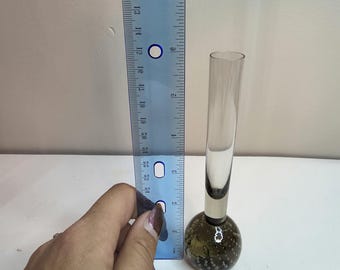 May include: A clear glass bud vase with a round, bubble-filled base and a slender, cylindrical neck. The vase is approximately 6 inches tall. The base is a dark olive green color. A blue ruler is next to the vase.