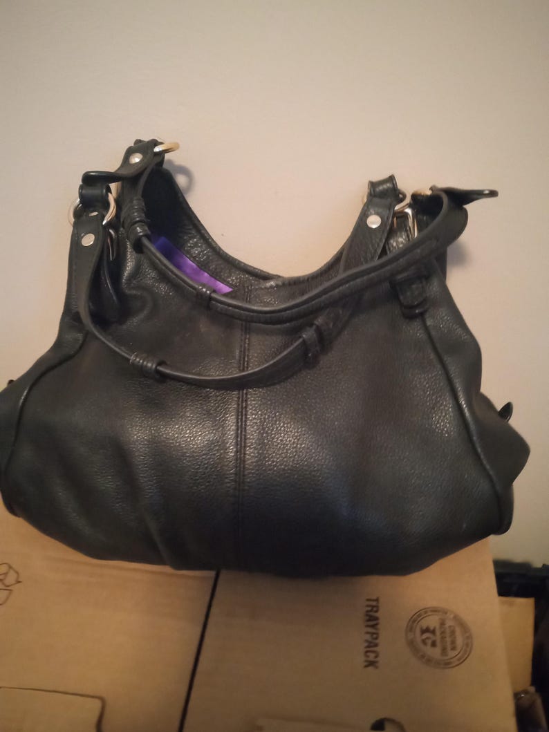 May include: Black leather hobo bag with a curved shape and a single shoulder strap. The bag has gold-colored hardware and a textured surface. The interior lining is purple. The bag is sitting on a cardboard box.