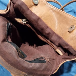 May include: A brown leather handbag with a zippered pocket and a brown fabric lining. The bag has a metal tag that says "VALENTINO".