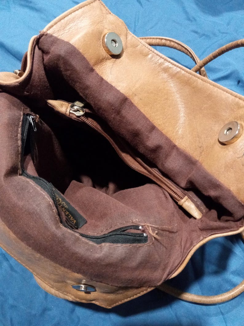May include: A brown leather handbag with a zippered pocket and a brown fabric lining. The bag has a metal tag that says "VALENTINO".