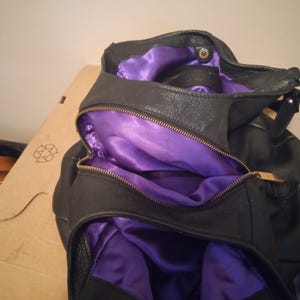 May include: Black leather handbag with a purple satin lining. The bag has a zipper closure and a silver-colored metal ring. The interior is visible, showing the contrast between the black exterior and the vibrant purple lining.