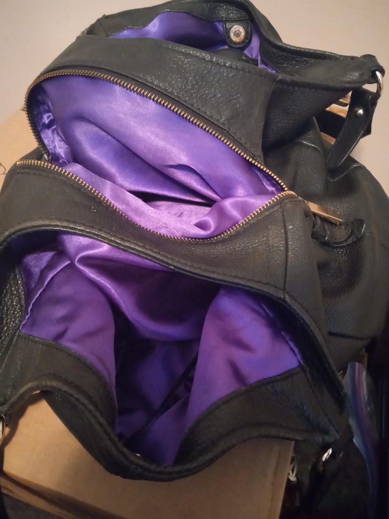 May include: Black handbag with a purple satin lining. The bag is open, revealing the interior. The zipper is gold-colored. The bag appears to be made of leather or a similar material.