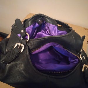 May include: Black leather handbag with silver-tone buckles and a purple satin interior. The bag has a zippered pocket and a magnetic snap closure. The bag is open, revealing the interior.