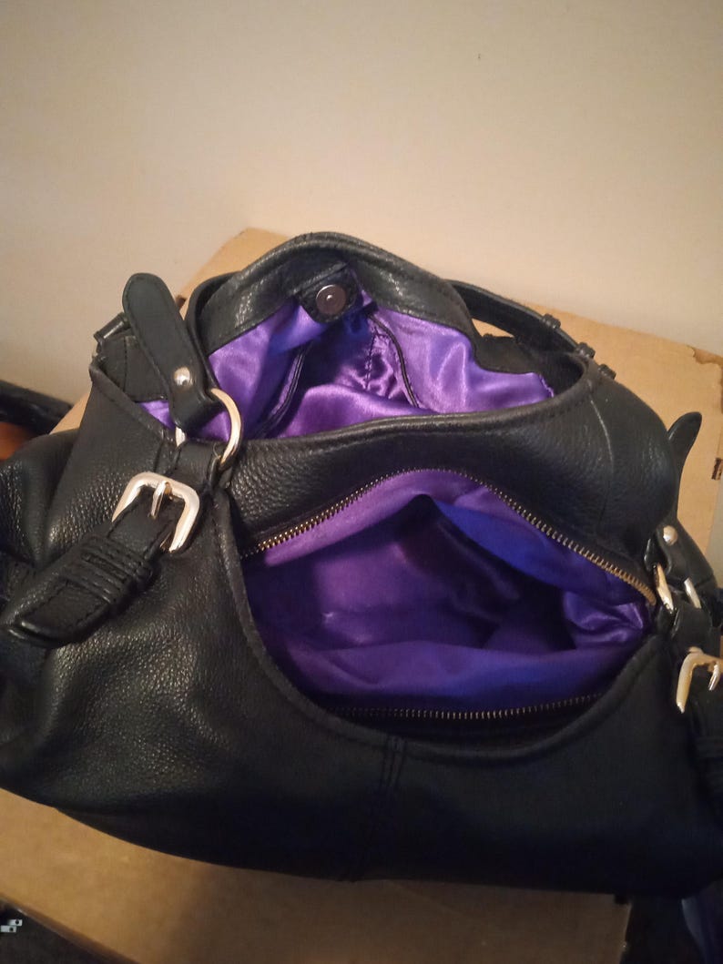 May include: Black leather handbag with silver-tone buckles and a purple satin interior. The bag has a zippered pocket and a magnetic snap closure. The bag is open, revealing the interior.