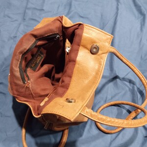 May include: A tan leather handbag with a brown fabric lining. The bag has a zippered pocket and a magnetic closure. The bag has two straps, one for carrying on the shoulder and one for carrying by hand.
