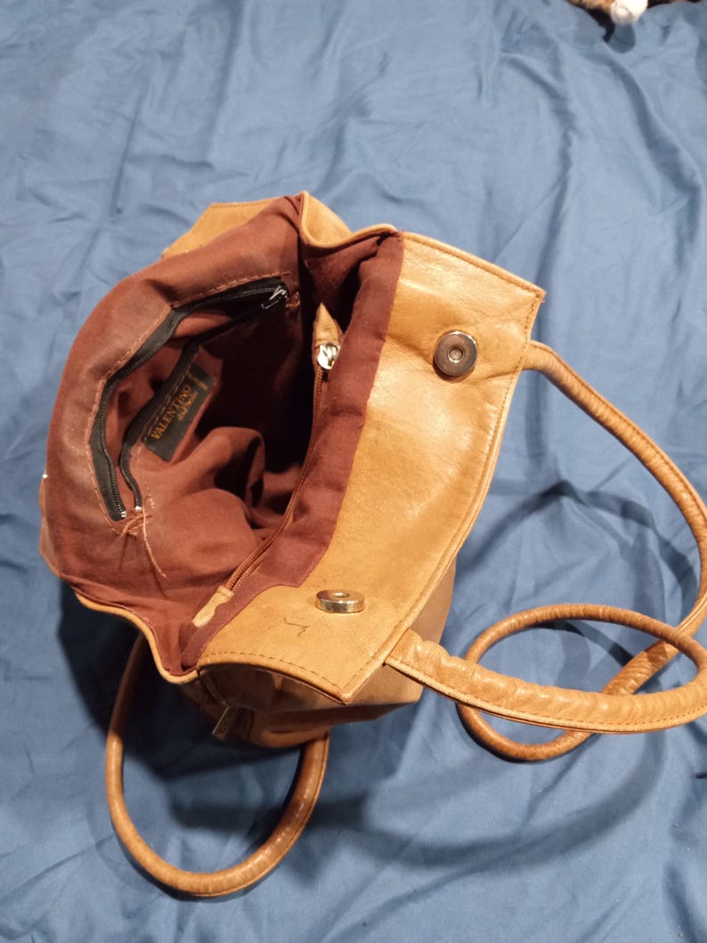 May include: A tan leather handbag with a brown fabric lining. The bag has a zippered pocket and a magnetic closure. The bag has two straps, one for carrying on the shoulder and one for carrying by hand.