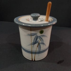 May include: A white ceramic honey pot with a blue and brown bamboo design. The pot has a lid and a wooden honey dipper.