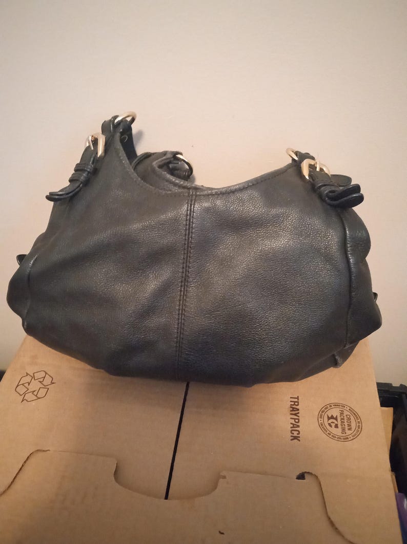 May include: Black leather hobo bag with gold-tone hardware. The bag features a curved shape, a single shoulder strap, and decorative details near the strap attachments. The bag is sitting on a cardboard box.