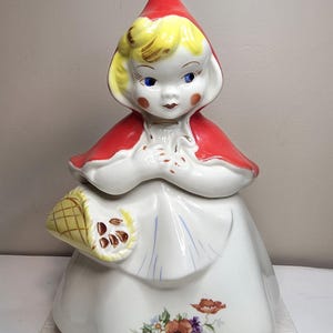 May include: A ceramic cookie jar shaped like a figure wearing a bright red hood and cape over a white dress. The figure has blonde hair, blue eyes, and rosy cheeks. A yellow basket is held at the side, and a floral pattern decorates the skirt.