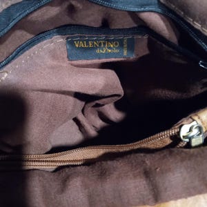 May include: The inside of a brown leather bag with a zipper closure. The bag has a brown fabric lining and a black label with the text "VALENTINO di Polo" in gold lettering.