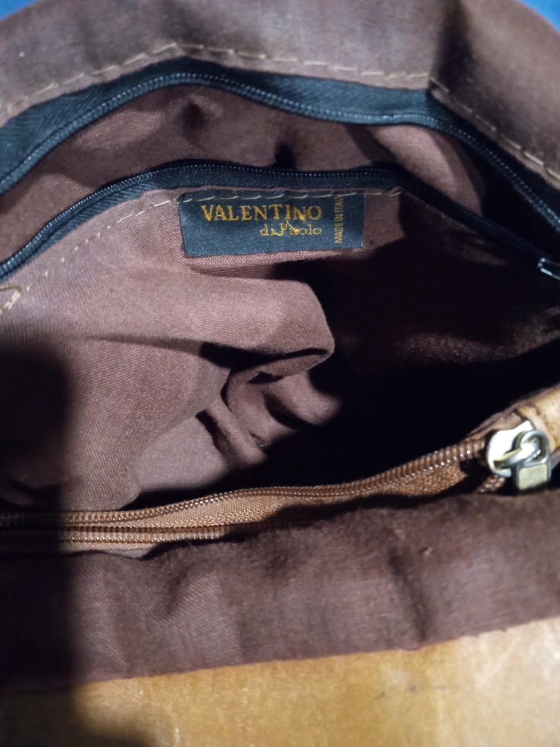 May include: The inside of a brown leather bag with a zipper closure. The bag has a brown fabric lining and a black label with the text "VALENTINO di Polo" in gold lettering.