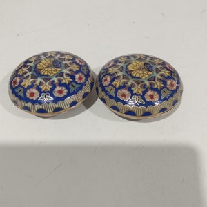Op de afbeelding: Twee ronde, decoratieve dozen met ingewikkelde bloemmotieven in tinten blauw, geel, roze en groen. De dozen hebben een gouden rand en zijn gemaakt van emaille.