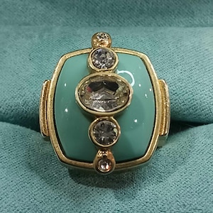 May include: A turquoise ring with a gold setting and five clear rhinestones. The ring has a square shape with rounded corners.