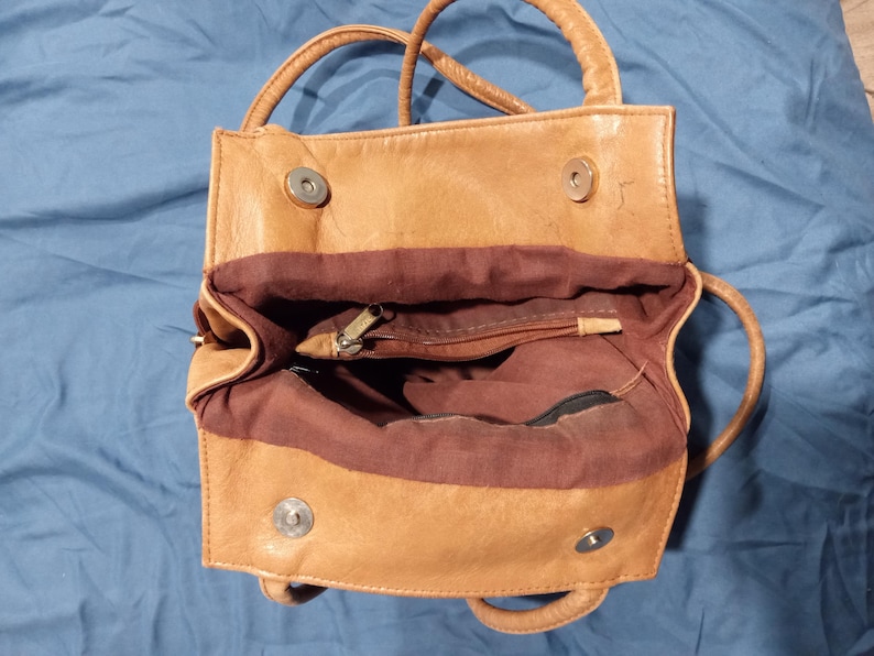 May include: A tan leather handbag with a brown fabric lining. The bag has two top handles and a zipper closure.