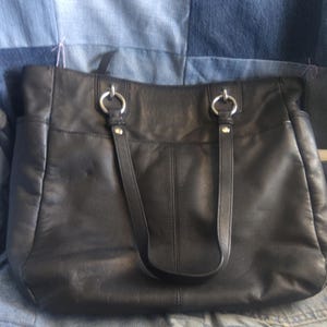 May include: A black leather tote bag featuring two shoulder straps attached with silver-toned metal rings and rivets. The bag has a rectangular shape with a central vertical seam and side pockets, showcasing a smooth texture and a classic design.