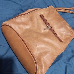 May include: A brown leather tote bag with a zippered pocket and a silver zipper pull. The bag has a brown leather strap.