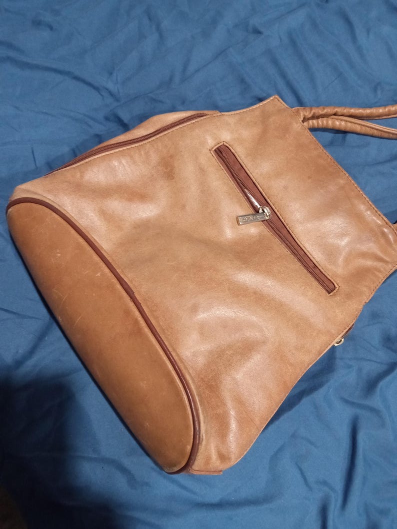 May include: A brown leather tote bag with a zippered pocket and a silver zipper pull. The bag has a brown leather strap.