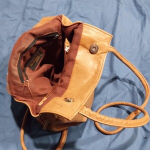 May include: A tan leather handbag with a brown fabric lining. The bag has a zipper closure and a snap closure. The bag has a brand label that reads "VALENTINO" inside.