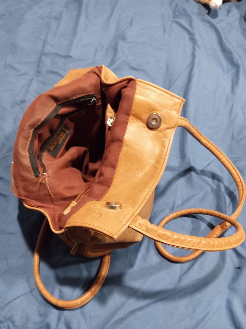 May include: A tan leather handbag with a brown fabric lining. The bag has a zipper closure and a snap closure. The bag has a brand label that reads "VALENTINO" inside.
