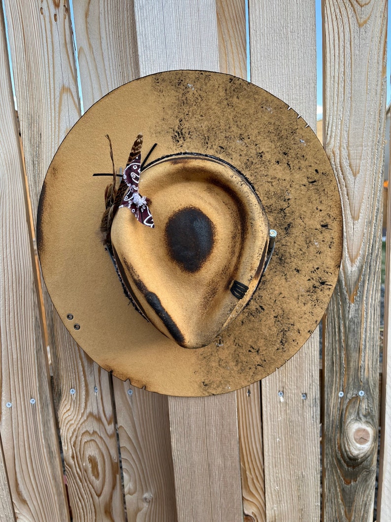 K&ouml;nnte beinhalten: Ein brauner Cowboyhut mit einem abgenutzten und verwitterten Aussehen. Der Hut hat ein braunes Band mit einem kleinen St&uuml;ck Stoff daran befestigt. Der Hut h&auml;ngt an einem Holzzaun.
