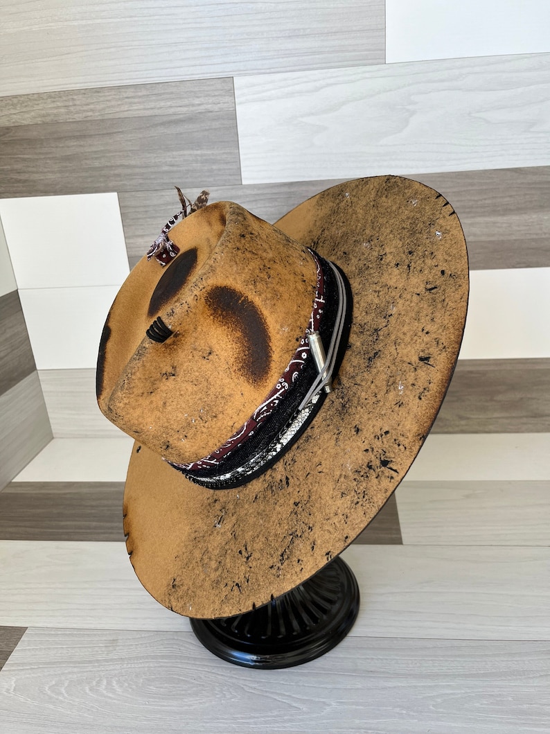K&ouml;nnte beinhalten: Ein brauner Filzhut im Cowboy-Stil mit einem abgenutzten Look, einem schwarzen und wei&szlig;en Bandana-Band und einer silbernen Schnalle. Der Hut wird auf einem schwarzen St&auml;nder pr&auml;sentiert.
