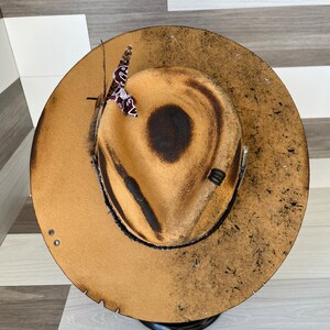 K&ouml;nnte beinhalten: Ein brauner Filzhut im Cowboy-Stil mit abgenutzter Oberfl&auml;che. Der Hut hat ein schwarzes Band und eine kleine braune Feder mit einem rot-wei&szlig;em Bandana.