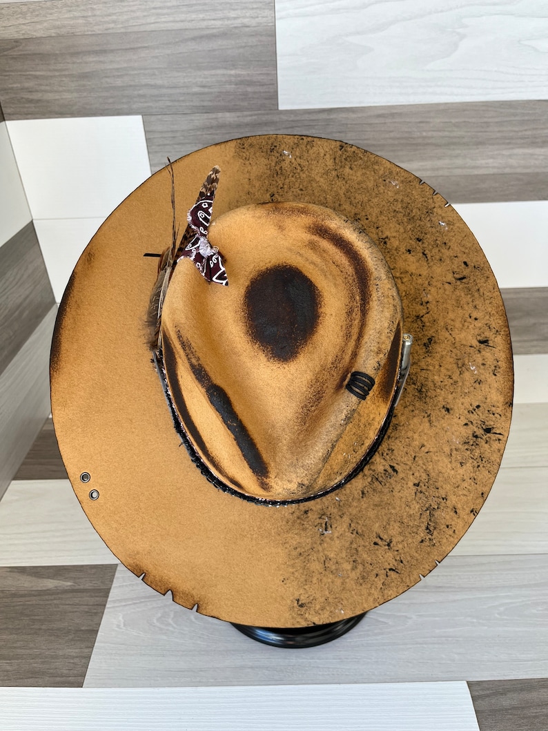 K&ouml;nnte beinhalten: Ein brauner Filzhut im Cowboy-Stil mit abgenutzter Oberfl&auml;che. Der Hut hat ein schwarzes Band und eine kleine braune Feder mit einem rot-wei&szlig;em Bandana.