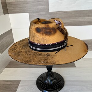 K&ouml;nnte beinhalten: Ein brauner Filzhut im Cowboy-Stil mit einem schwarzen Band und einem braunen und wei&szlig;en Bandana. Der Hut hat einen abgenutzten Look mit braunen Flecken und einem Federschmuck.