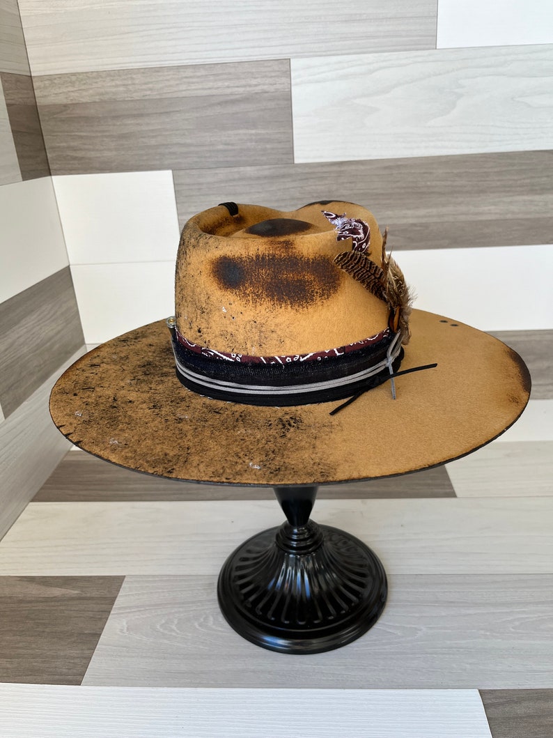 K&ouml;nnte beinhalten: Ein brauner Filzhut im Cowboy-Stil mit einem schwarzen Band und einem braunen und wei&szlig;en Bandana. Der Hut hat einen abgenutzten Look mit braunen Flecken und einem Federschmuck.