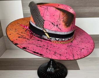 Pode incluir: Um chapéu de cowboy rosa e laranja com respingos de tinta preta e um detalhe de pena. O chapéu está em um suporte preto.