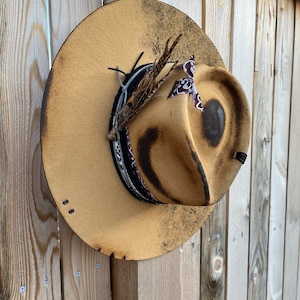 K&ouml;nnte beinhalten: Ein brauner Filzhut im Cowboy-Stil mit einem schwarzen Band und einer braunen Feder. Der Hut hat einen abgenutzten Look mit ein paar dunklen Flecken und einem kleinen Riss am Rand.