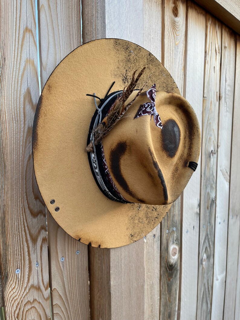K&ouml;nnte beinhalten: Ein brauner Filzhut im Cowboy-Stil mit einem schwarzen Band und einer braunen Feder. Der Hut hat einen abgenutzten Look mit ein paar dunklen Flecken und einem kleinen Riss am Rand.