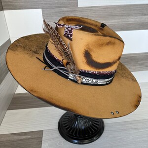 K&ouml;nnte beinhalten: Ein brauner Filzhut im Cowboy-Stil mit einem abgenutzten Look. Der Hut hat ein schwarzes und wei&szlig;es Bandana-Band mit einem Federschmuck.
