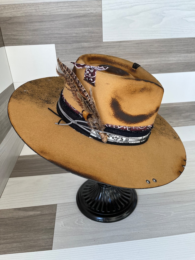 K&ouml;nnte beinhalten: Ein brauner Filzhut im Cowboy-Stil mit einem abgenutzten Look. Der Hut hat ein schwarzes und wei&szlig;es Bandana-Band mit einem Federschmuck.