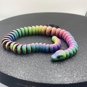 May include: A multi-coloured, articulated snake toy. The toy is made of connected, rounded segments, with a small, detailed head. The snake is resting on a dark, circular surface.