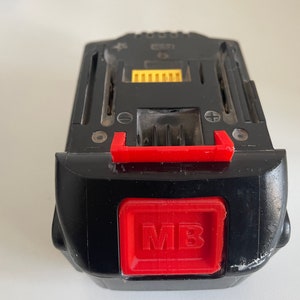 May include: Black power tool battery with a red handle and a red square button with the letters "MB". The battery has a yellow connector and is designed for power tools.