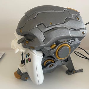 May include: A grey and gold skull-shaped gaming controller stand. It holds a white controller with colourful buttons. The design has a detailed, mechanical aesthetic with a skull-like face and a sturdy base.