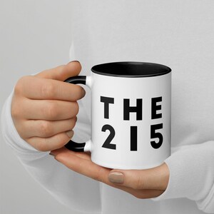 May include: White ceramic mug with a black rim and handle. The mug has black text that reads "THE 215".