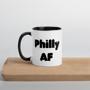 May include: White ceramic mug with a black interior and handle. The mug features the text "Philly AF" in bold, black letters. The mug is sitting on a wooden surface.
