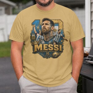 May include: A tan crew neck t-shirt featuring a graphic print of Lionel Messi. The design includes a large portrait of the player, two smaller action shots, a gold crown, a soccer ball, the text "MESSI", "TALENTO SEM LIMITES, LENDARIO PARA SEMPRE", and "10".