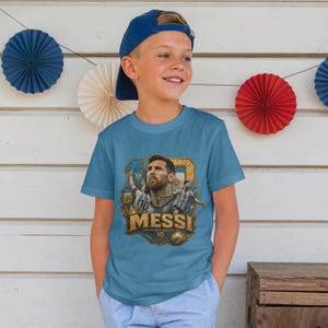 May include: A blue short-sleeved t-shirt featuring a graphic print of Lionel Messi. The design includes the name MESSI in large gold letters, the number 10, a soccer ball, and multiple images of the athlete in an Argentina soccer jersey.
