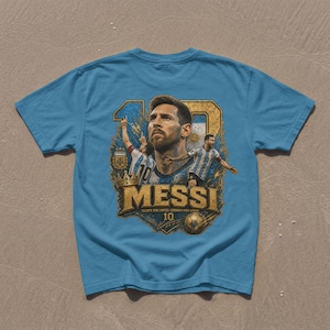 May include: A light blue t-shirt featuring a graphic print of Lionel Messi. The design includes the number 10, a portrait of the player, action shots in an Argentina jersey, a gold crown, a soccer ball, and the text MESSI, TALENTO SEM LIMITES, LEND&Aacute;RIO PARA SEMPRE.
