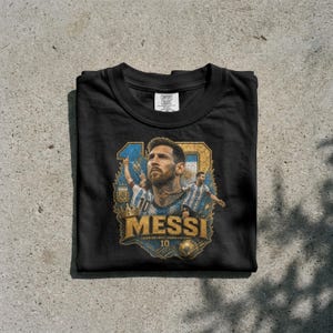 May include: A black crew neck t-shirt featuring a graphic print of Lionel Messi. The design includes a large portrait of Messi, two smaller action figures in Argentina jerseys, a gold crown, a soccer ball, and the text "MESSI" and "10".