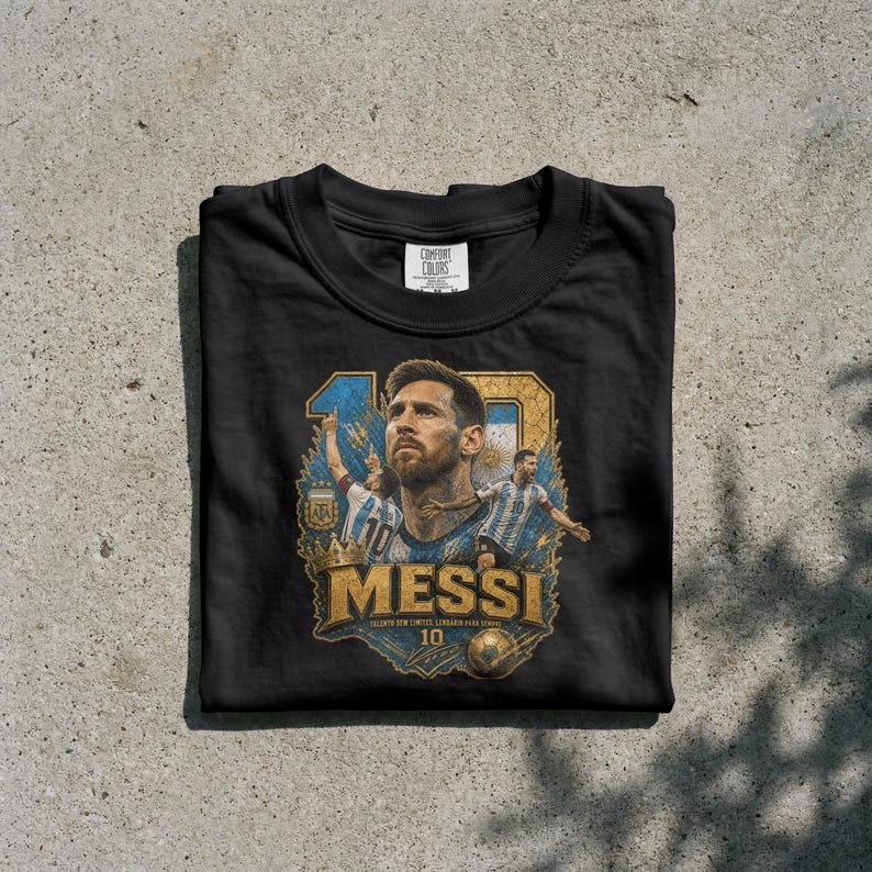 May include: A black crew neck t-shirt featuring a graphic print of Lionel Messi. The design includes a large portrait of Messi, two smaller action figures in Argentina jerseys, a gold crown, a soccer ball, and the text "MESSI" and "10".