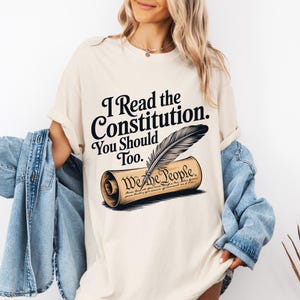 May include: A cream-coloured short-sleeve t-shirt featuring the black text "I Read the Constitution. You Should Too." above an illustration of a quill pen resting on a rolled parchment document with the words "We the People" printed on it.