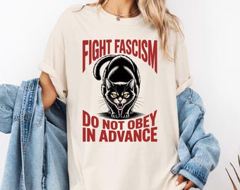 May include: Cream-colored t-shirt featuring a black cat illustration with the text "FIGHT FASCISM" above and "DO NOT OBEY IN ADVANCE" below. The cat has yellow eyes and is in a stalking pose. The text is in red.
