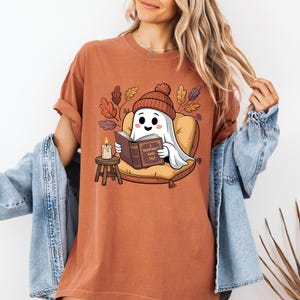 May include: Rust-colored t-shirt featuring a cartoon ghost wearing a knit cap and reading a book. The ghost is seated on a pillow with autumn leaves in the background. The book reads "A Hauntingly Good Tale."