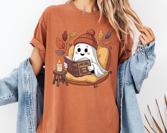 Op de afbeelding: Roestkleurig T-shirt met een cartoongeest met een gebreide muts en een boek lezend. De geest zit op een kussen met herfstbladeren op de achtergrond. Het boek luidt "A Hauntingly Good Tale".