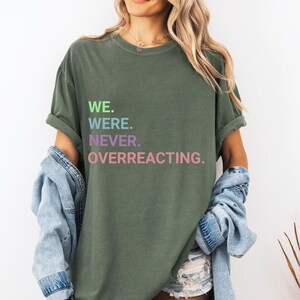Pode incluir: Camiseta verde-oliva com o texto "WE. WERE. NEVER. OVERREACTING." em verde, azul, roxo e rosa. A camiseta tem mangas enroladas e é combinada com uma jaqueta jeans.